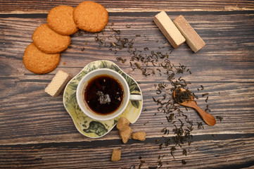 A Cup of black tea, tea leaves, pieces of brown sugar, oatmeal cookies, waffles on a wooden background. Close up.