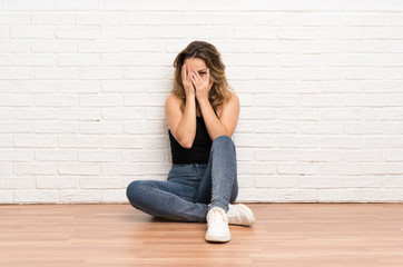Young blonde woman sitting on the floor covering eyes and looking through fingers