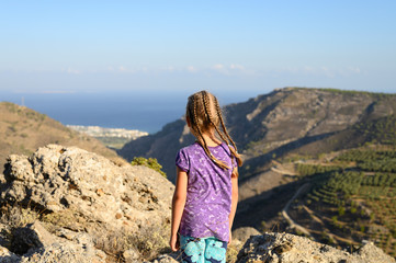 A little girl admires the seascape. Family with kids hiking in Greece mountains. Beautiful landscape with hills, sea and sky with low clouds. Outdoor activity in the nature for parents and children