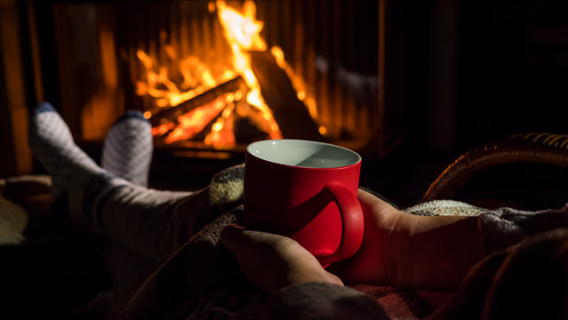 Woman with a red cup of tea is relaxing by the fireplace