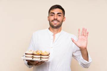 Handsome man holding muffin cake over isolated background saluting with hand with happy expression