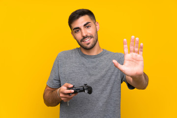 Fototapeta na wymiar Young handsome man playing with a video game controller over isolated yellow background saluting with hand with happy expression