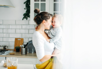 Young woman mom with baby girl on hands havinh fun and cooking breakfast on bright kitchen at home
