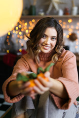 The concept of Christmas home shooting. Beautiful attractive cute brunette curly girl in warm home clothes laughs and poses against the background of Christmas dark kitchen, garlands and bokeh.