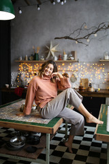 The concept of Christmas home shooting. Beautiful attractive cute brunette curly girl in warm home clothes laughs and poses against the background of Christmas dark kitchen, garlands and bokeh.