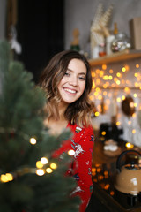 An attractive short-haired girl with a beautiful smile in a homemade Christmas red pajama jumpsuit smiles, laughs and indulges in the Christmas kitchen on a background of bokeh and garlands.