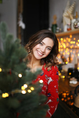 An attractive short-haired girl with a beautiful smile in a homemade Christmas red pajama jumpsuit smiles, laughs and indulges in the Christmas kitchen on a background of bokeh and garlands.