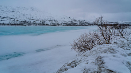 lac gel&eacute; dans un paysage enneig&eacute; et de la glace bleue avec des buissons sans feuilles au premier plan et des montagnes au fond sous un ciel gris sans soleil