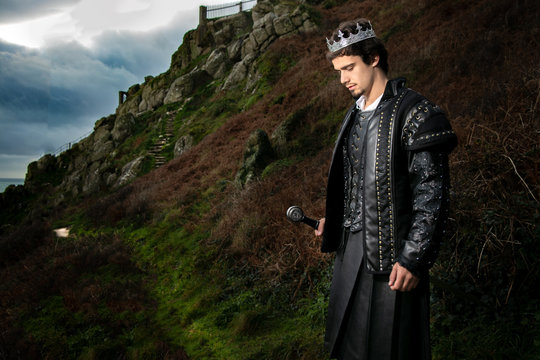 Handsome king with sword stands in contemplation with hill and parts of keep in the background