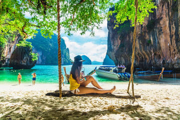 Traveler woman in bikini sitting on swing using tablet on tropical beach Lao Lading island Krabi,...