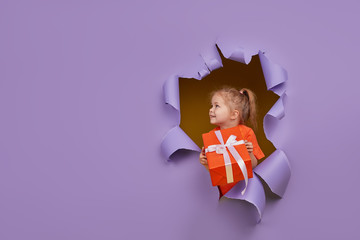 Cute little child girl breaks through a colored purple paper wall with gift box in hands. Toddler...