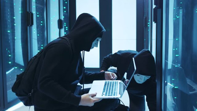 Confident internet criminals sneaking into database connecting laptop for coding. Group of masked hackers in hoodies stealing data center confidential information on cybercrime.