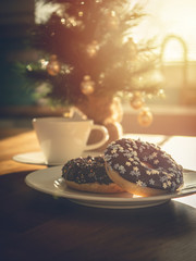 Chocolate donuts sprinkled with colorful stars on the table. Coffee and Christmas decorations in the background. Donut on Christmas table. Chocolate donut at sunrise. Coffee and donut.