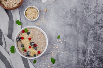 Oatmeal in a bowl with banana, frozen berries, almond petals, mint leaves and honey on a gray stone background. Tasty and healthy breakfast. Copy space.