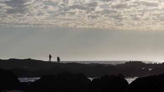 Beautiful silhouette of romantic couple walking on sea rocks, exploring. Ocean waves breaking around them. Sunset clouds. Sea and ocean sound. Sunlight on ocean. Summer vacation and holiday solitude. 