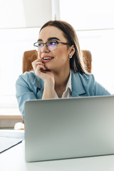 Young beautiful businesswoman in glasses working on laptop and keeping hand on chin while sitting at her working place.