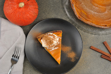 A piece of pumpkin pie on a black plate.  The cake is served with whipped cream.  In the frame, cinnamon sticks, pumpkin, pie, fork.  View from above.