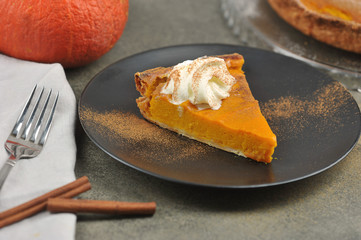 A piece of pumpkin pie on a black plate. The cake is served with whipped cream. The concept of a festive treat of the autumn harvest.  Close-up.  In the frame, cinnamon sticks, pumpkin, pie. 