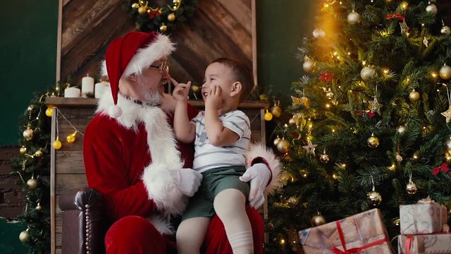 Little boy sitting on Santa Claus lap listening to his story, next to the Christmas tree in room with festive interior. Shooting in slow motion.