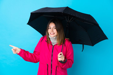 Young woman holding an umbrella over isolated blue wall surprised and pointing side