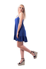 Fresh playful young blond hair surprised woman in blue dress or nightie posing with leg raised looks behind. Full body isolated on white background. 