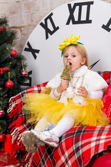 A child, a little girl sitting near a Christmas tree and eating a delicious lollipop in the form of a Christmas tree.