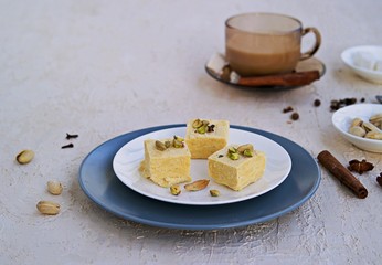 Indian sweets with pistachios, almonds and cardamom on a white plate on a light concrete...