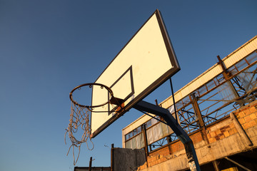 Broken basketball net dangling from a hoop attached to the white backboard. Defunct playground in recreational area, next to an abandoned and demolished industrial hall. Sunny day and clear blue sky.