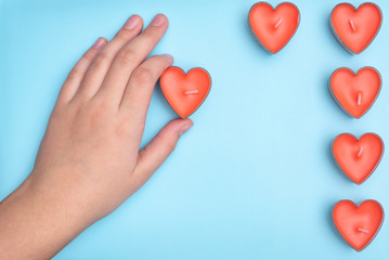 Fototapeta premium Flat lay a red candle in the form of a heart in children's hands and the remaining candles are located nearby isolated on light blue backgound. Wedding card ideas and Valentines Day