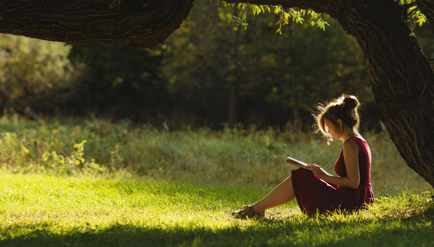 sunny portrait of a beautiful girl sitting on green glade under an arch of tree branches with book, woman reading novel on nature, concept hobby and lifestyle