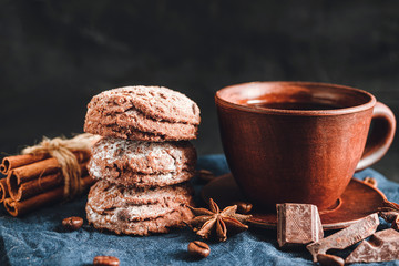 Homemade bakery, chocolate cookies with powdered sugar, brown cup with coffee, cinnamon sticks, star anise on blue napkin, dark background