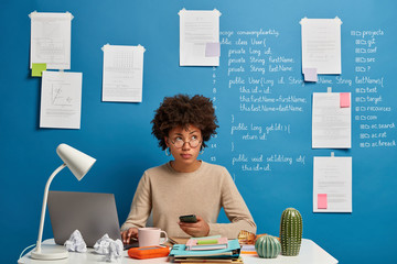 Busy curly Afro woman works from home, uses laptop and smartphone at workplace, checks newsfeed,...