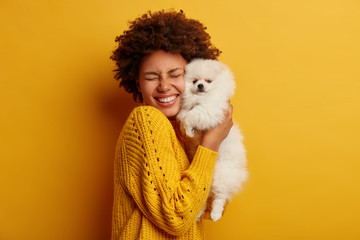 Pleased happy Afro girl gets lovely puppy, plays and embraces four legged friend with love, stands...