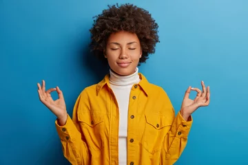 Fotobehang Relieved calm dark skinned woman closes eyes, makes mudra gesture, dressed in yellow shirt, feels relaxed, poses against blue background, releases stress, stands pecefully indoor in lotus pose © Wayhome Studio