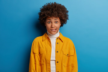 Fototapeta na wymiar Portrait of worried dark skinned woman bites lips and looks surprisingly at camera, dressed in white turtleneck and yellow shirt, being nervous, isolated on blue, hesitates while wants ask question