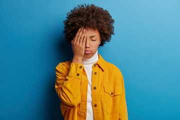 Fototapeta na wymiar Tired fed up woman covers half of face, keeps eyes closed, sighs from tiredness, feels exhausted to work without rest, poses in studio against blue wall, wears stylish clothing, wants to sleep