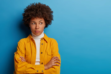 Beautiful ethnic woman gazes thoughtfully on right side, keeps hands crossed over chest, wears yellow shirt, poses against blue background, faces hard choice, solves puzzle, makes up plan in mind