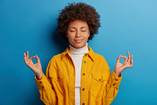 Relieved calm dark skinned woman closes eyes, makes mudra gesture, dressed in yellow shirt, feels relaxed, poses against blue background, releases stress, stands pecefully indoor in lotus pose