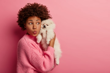 Studio shot of funny curly haired woman keeps lips folded, enjoys free time with cute miniature breed puppy, keeps spitz dog closely near face, wears oversized jumper, relaxes together with animal