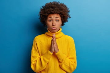 Fototapeta na wymiar Image of sad hopeful woman with curly hairdo holds hands pressed together as sign of hope, makes praying gesture, needs support and help, wears yellow jumper, wishes and believes in miracle.