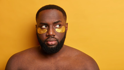 Serious Afro man has healthy dark skin, collagen patches under eyes, stands naked against yellow background, has anti aging procedure, looks thoughtfully aside. Skin care and cosmetology concept