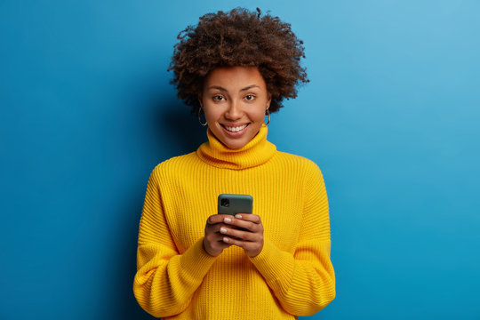 Photo Of Dark Skinned Lady Enjoys Distant Communication, Uses Mobile Phone, Surfes Fast Unlimited Internet, Has Pleasant Smile, Dressed In Yellow Sweater, Makes Shopping Online, Isolated On Blue