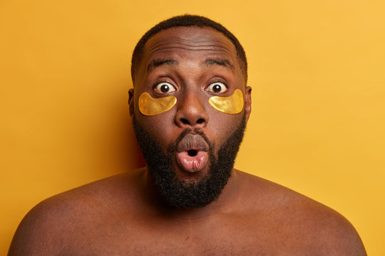 Cosmetology for men. Close up shot of dark skinned man looks with surprised face expression, wears undereye patches for reducing puffiness, has thick stubble, isolated over yellow background