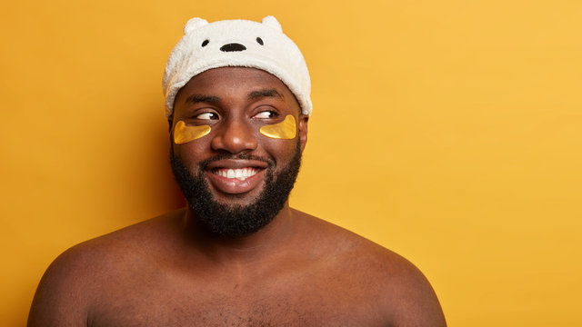 Smiling Black Male Model Has Happy Expression, Looks Aside, Applies Golden Patches To Cure Swelling, Has Midlife Crisis, Wears Soft Funny Hat, Stands Bare Shoulders Against Bright Yellow Wall. Beauty