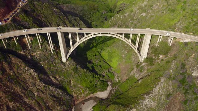 Aerial view of the picturesque arch bridge standing over the canyon among the beautiful hills covered with green grass. Cars move across the bridge. Famous landmark of USA under the cloudy sky. 4K