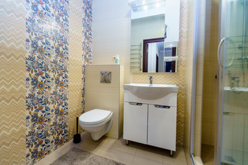 Bathroom in the apartment. Shower, sink with mirror, toilet. Square ceramic tiles in the form of yellow-red-blue floral patterns on the wall in the bathroom.