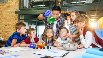 Pupils of primary school watch carefully for their teacher who shows interesting chemical experiments with colored liquids in glass flaks.