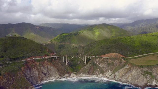 Breathtaking aerial footage of the mountain landscape and rocky coast washed by vibrant blue water of Pacific ocean. Drone camera moves away from the graceful arch bridge set close to the water. 4K
