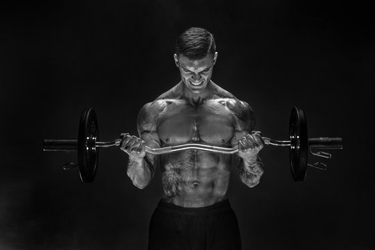 Studio portrait of topless bodybuilder performing biceps exercise with concentrated face over black background with smoke. Cutout. Very brawny guy bodybuilder.