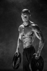 Fototapeta na wymiar Very brawny guy bodybuilder. Bodybuilder with dumbbells in his arms on dark background with smoke.
